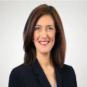 A woman with straight brown hair wearing a black blazer and light blue top, smiling at the camera against a plain white background.