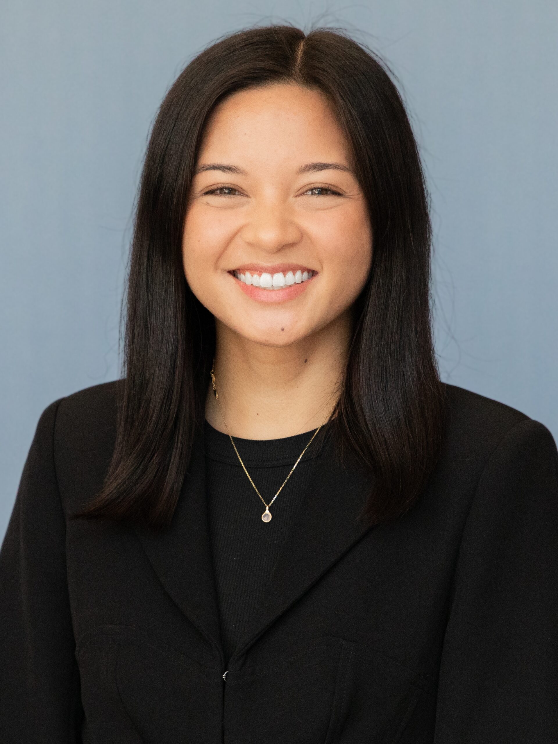 A woman with straight, shoulder-length dark hair, part of the D.C. Fellow Program, smiles at the camera. She is wearing a black blazer, a black top, and a gold necklace with a small pendant against a plain light blue background.