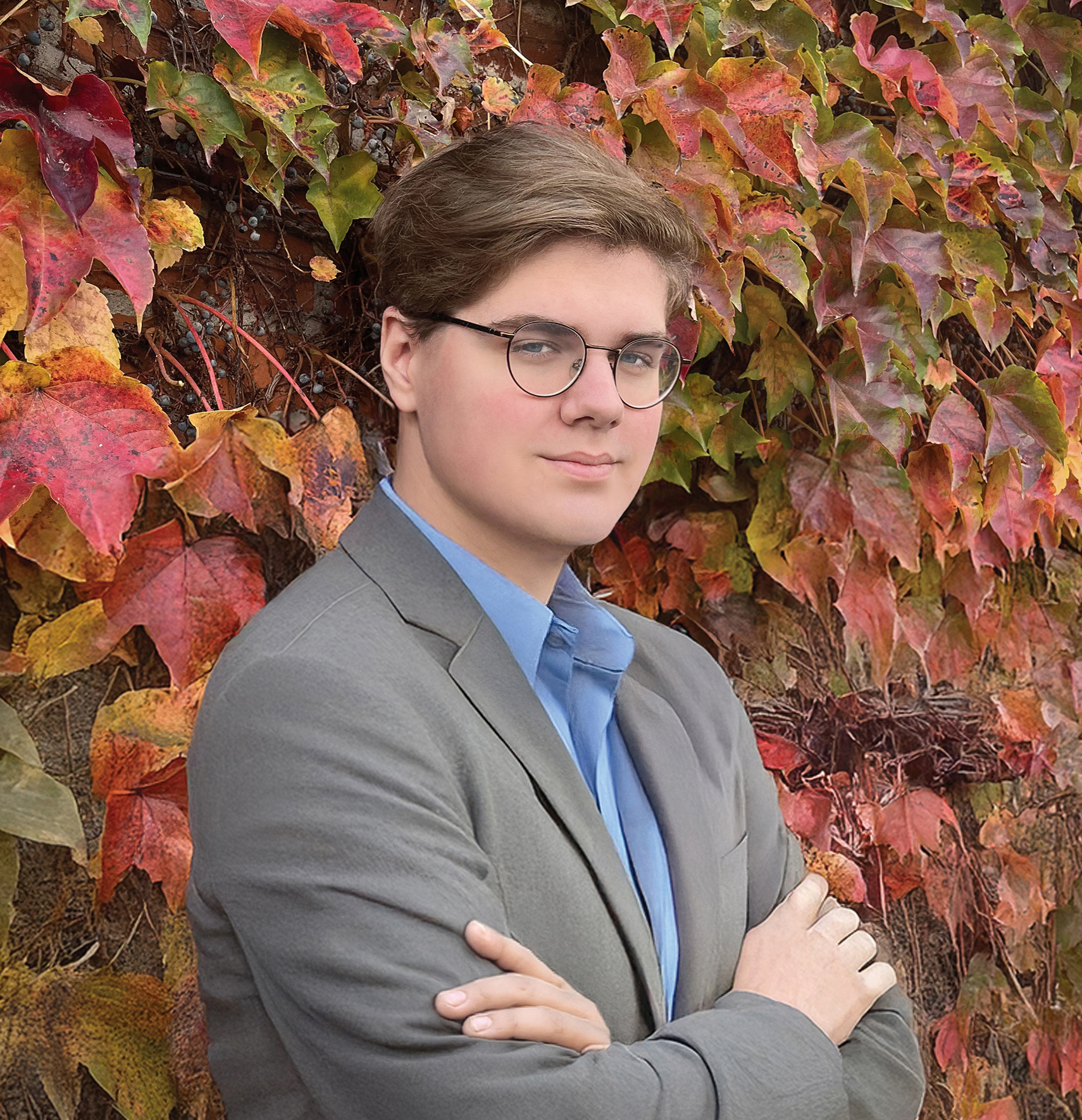 BrendenCunningham A person with glasses and light brown hair, wearing a gray blazer and blue shirt, stands with arms crossed in front of a wall covered in colorful autumn ivy leaves.