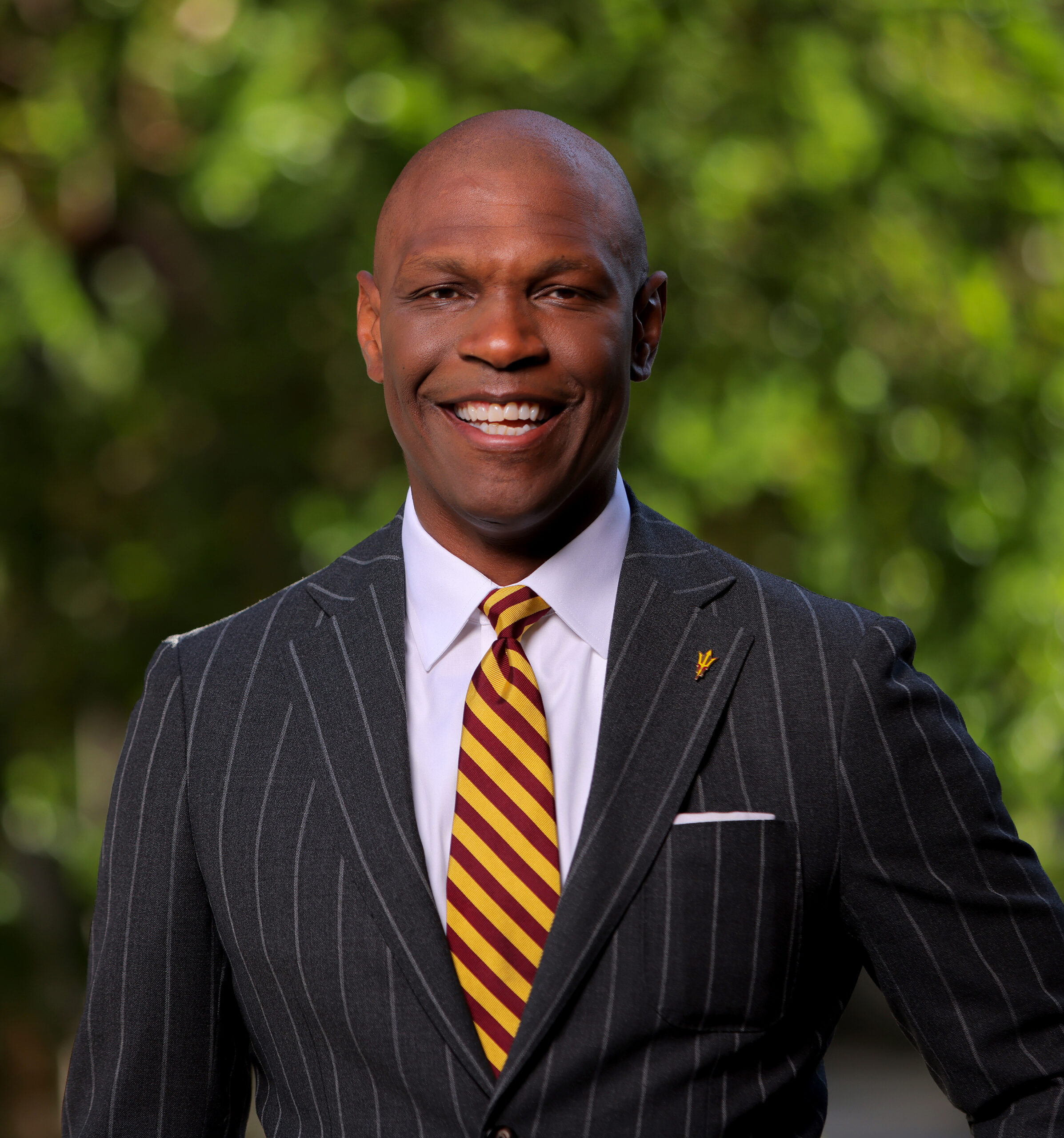 CBH Headshot A smiling man in a dark pinstripe suit and a maroon and gold striped tie stands outdoors in front of a leafy green background, representing his 501(c)(3) nonprofit organization with pride.