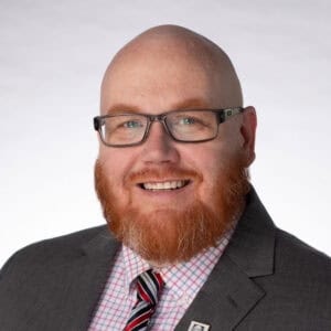 A bald man with a red beard and glasses smiles while wearing a gray suit, striped tie, and pink checkered shirt against a plain light background.