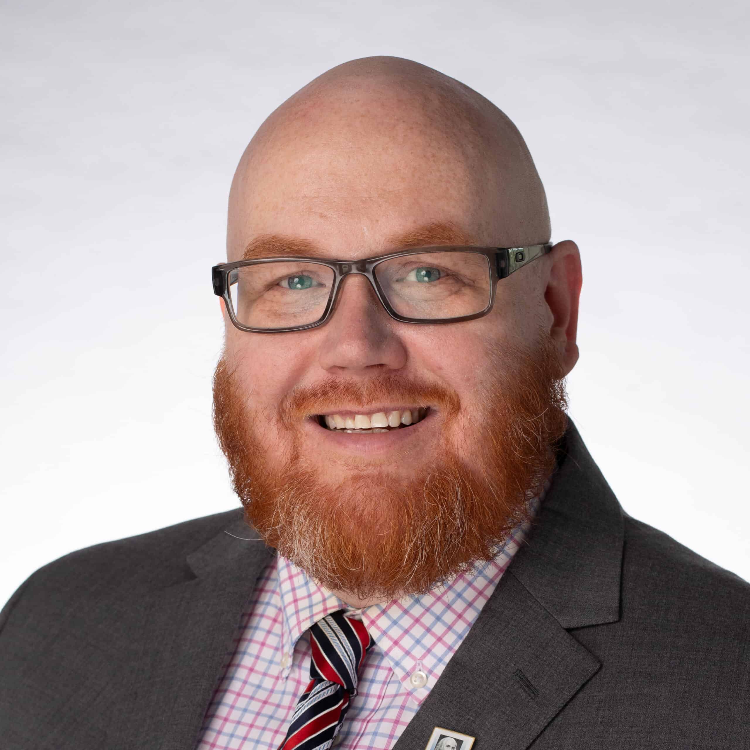 A bald man with a red beard and glasses smiles while wearing a gray suit, striped tie, and pink checkered shirt against a plain light background.