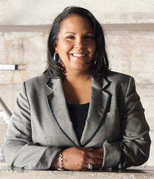 A person with long dark hair, wearing a grey blazer and a black top, is smiling while leaning against a concrete surface. The background is a muted, textured wall.