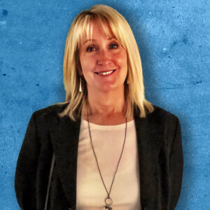 A person with blonde hair is smiling against a blue-textured background. They are wearing a white shirt, a black blazer, and a necklace.