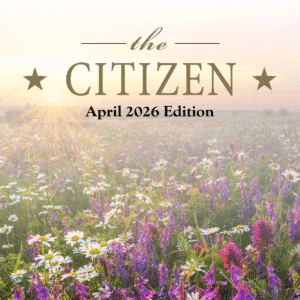 A sunlit field of wildflowers with text overlay: "The Citizen, April 2026 Edition." The scene captures a bright sunrise over colorful flowers under a soft, hazy sky, reflecting the vibrant spirit of The Citizen.