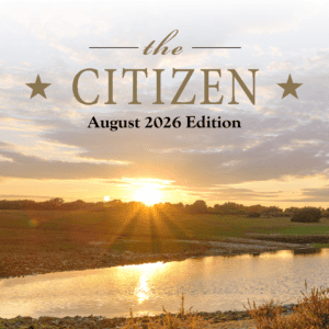 Sunset over a pond with grassy banks and trees. Text reads: “The Citizen August 2026 Edition” with two stars.