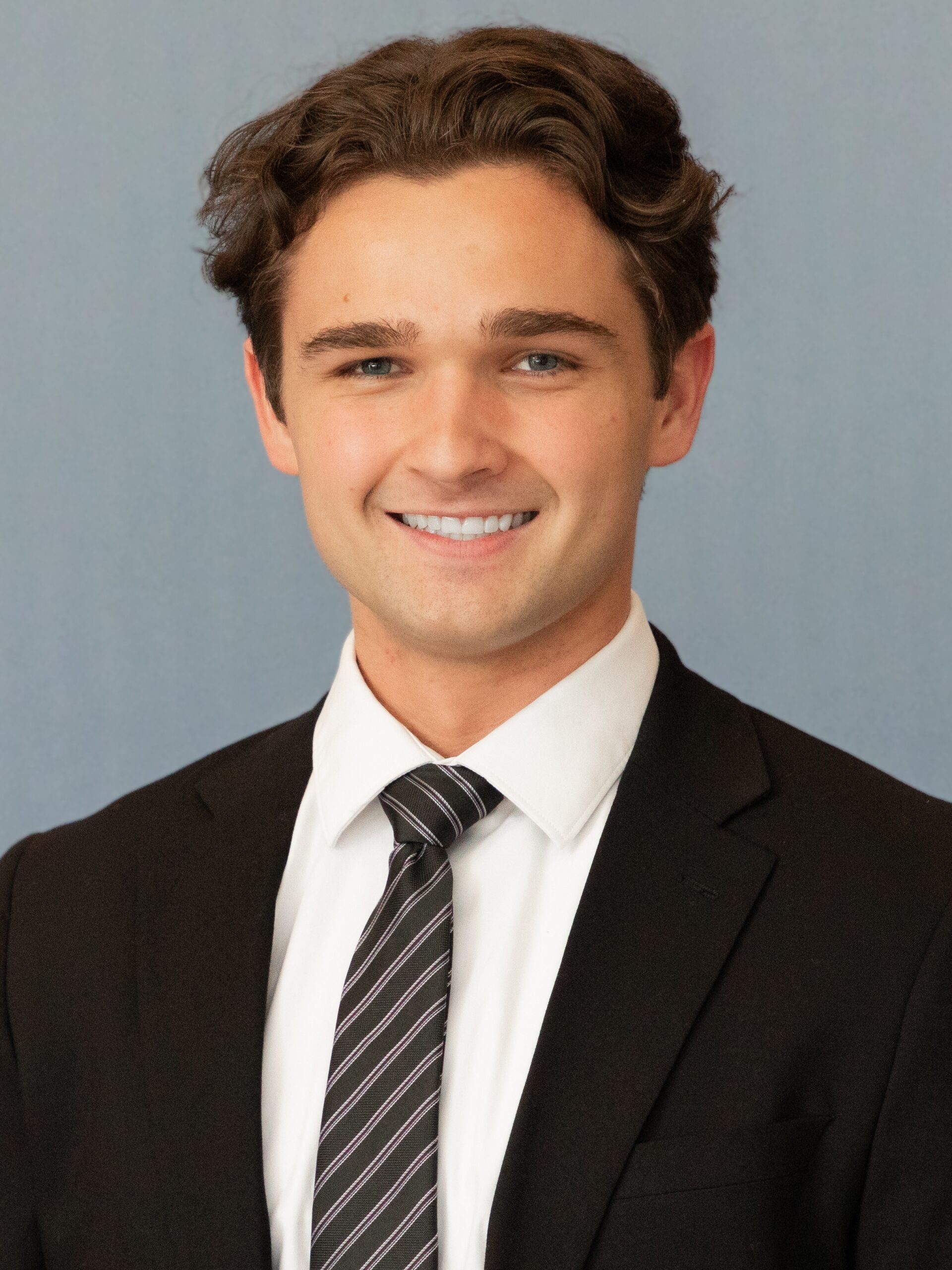 DallinHeadshot A young man with wavy brown hair is smiling, wearing a black suit, white dress shirt, and striped tie, standing against a plain light blue background—perfectly dressed for making Capital Connections.