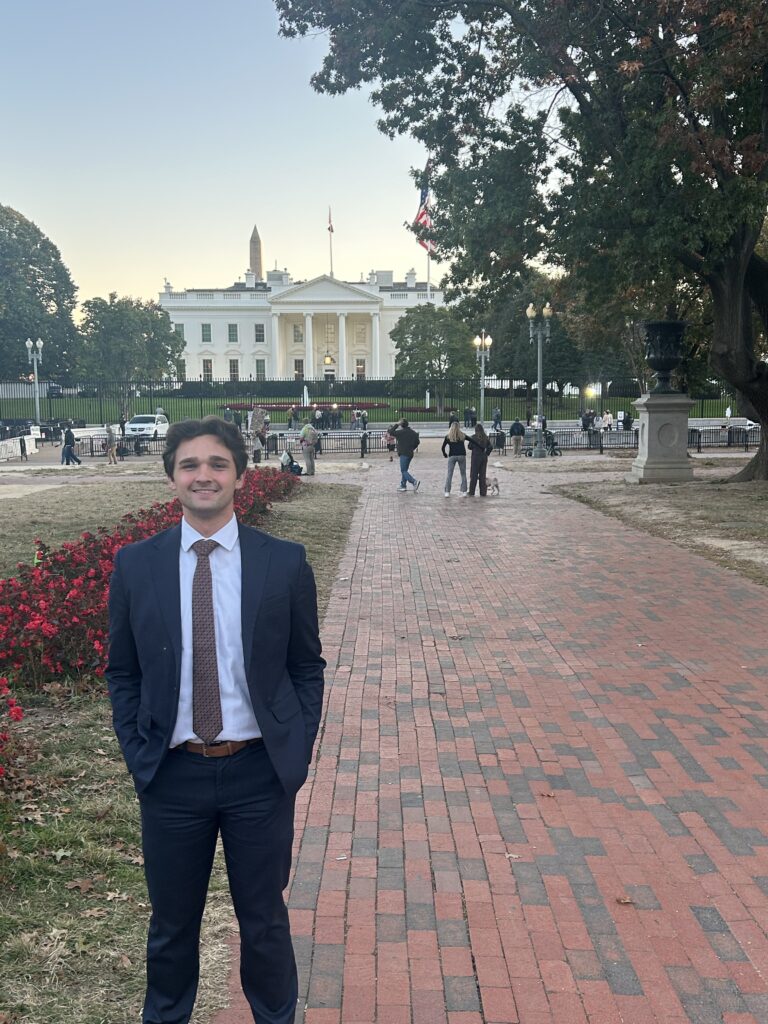 A man in a suit stands on a brick walkway with his hands in his pockets, smiling in front of the White House—an image that captures the essence of Capital Connections amidst people and trees in the background.