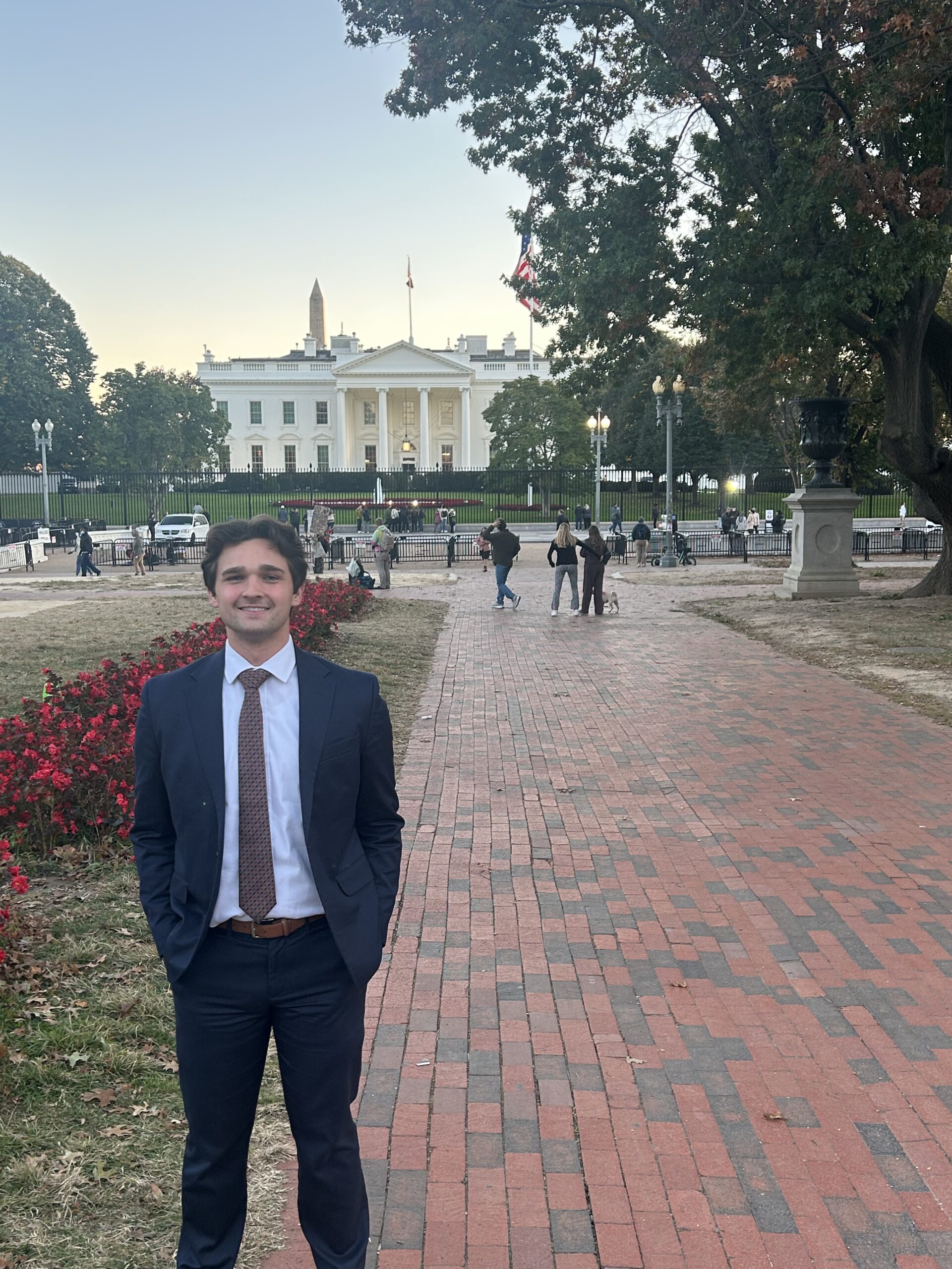 DallinInDC A man in a suit stands on a brick walkway with his hands in his pockets, smiling in front of the White House—an image that captures the essence of Capital Connections amidst people and trees in the background.