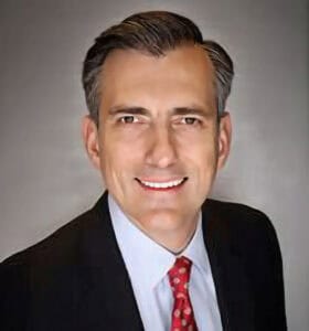 A man in a dark suit and red patterned tie smiles at the camera against a plain, gray background, as if posing for a history dinner event. He has short, neatly styled dark hair with gray highlights.