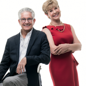 A smiling man in a dark blazer and white shirt sits on a stool, while a woman in a red dress stands beside him, leaning with her arms crossed. Both have short, light hair and exude the warmth of an old-fashioned history dinner set against a plain white background.