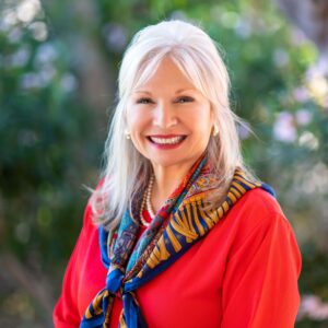 A smiling woman with long white hair wearing a red top and a colorful scarf stands outdoors. The background is blurred greenery.