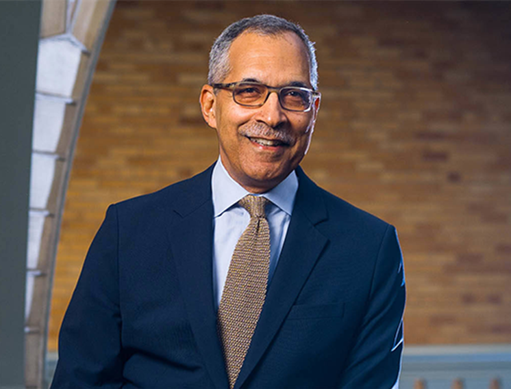 A smiling man in a navy suit, light blue shirt, and patterned tie stands in front of a brick wall. He has short gray hair and is wearing glasses.