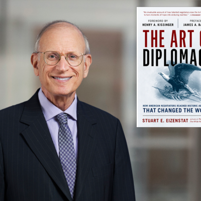 An older man in a suit and tie smiles at the camera. To his right is a book cover titled "The Art of Diplomacy" by Stuart E. Eizenstat, featuring an eagle and a globe illustration. The background is blurred.