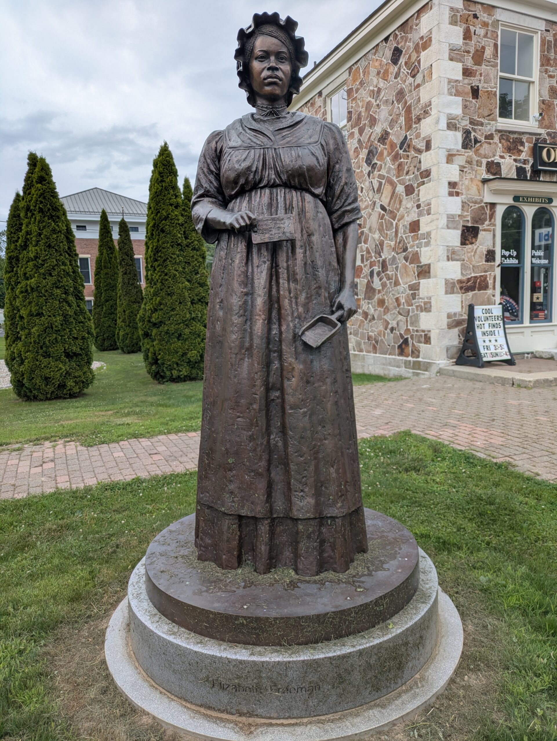 A bronze statue of a woman in a long dress holding a book stands on a round stone base outdoors, near a stone building and trimmed evergreen trees—a striking tribute often highlighted during Black History Month.