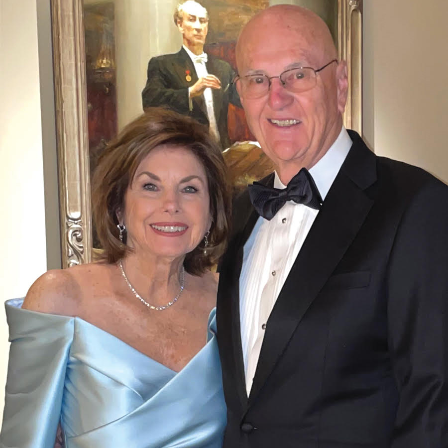 A smiling couple dressed formally attends a history dinner; the woman wears a light blue off-the-shoulder gown and the man sports a black tuxedo with a bow tie. They stand in front of a framed painting of a man in a suit.