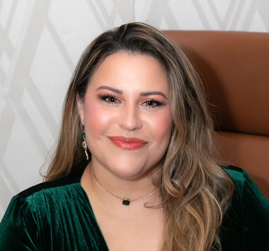 A woman with long, wavy blonde hair, wearing a green velvet top, gold earrings, and a necklace, smiles while sitting in front of a brown chair and patterned background.