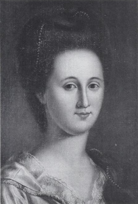 Black-and-white portrait of a woman with an elaborate hairstyle, wearing a dress with lace trim. In honor of Women's History Month, she looks directly at the viewer with a calm expression against a plain background.