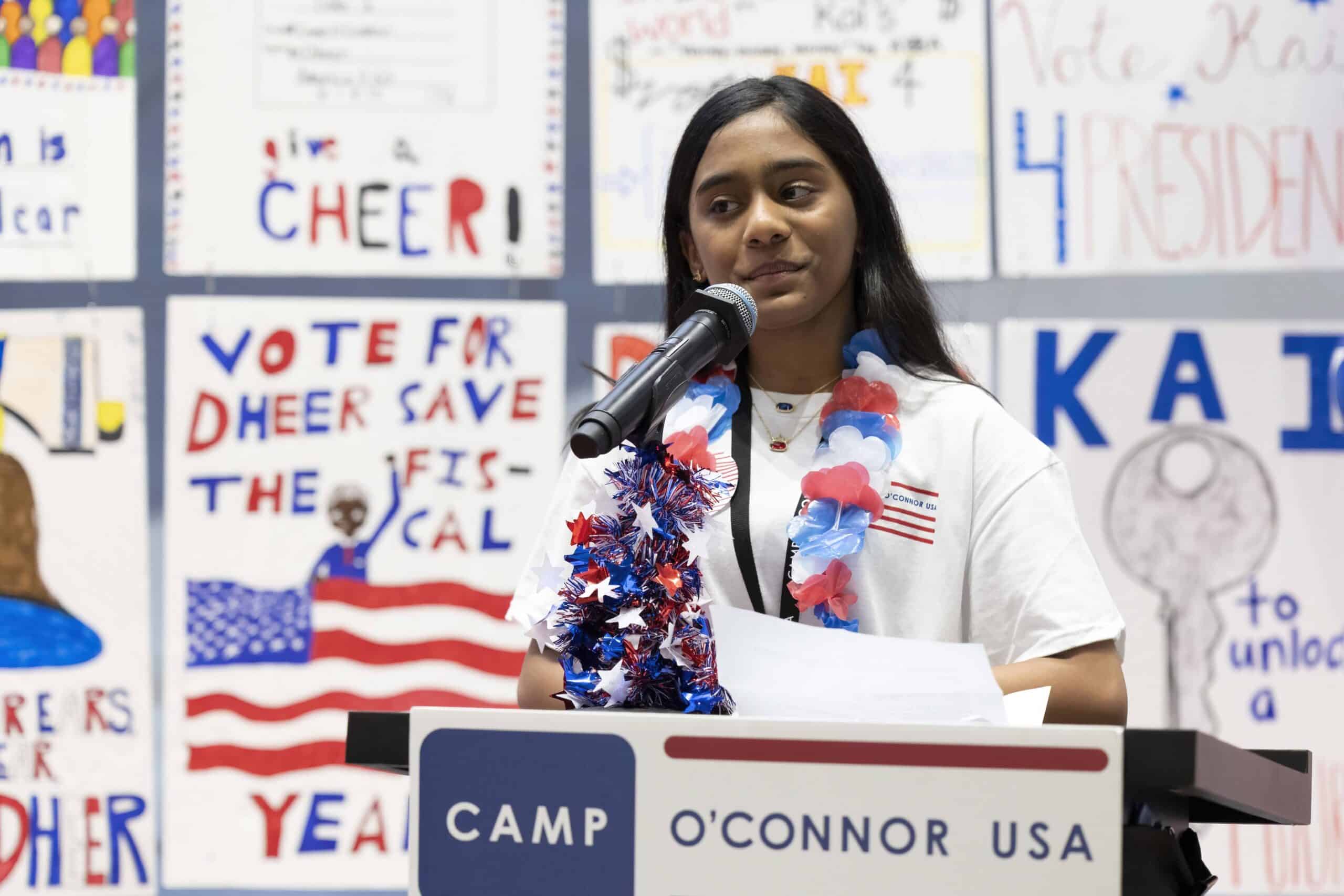 A young woman wearing a patriotic lei speaks at a podium labeled "Camp O’Connor USA," with campaign posters and an American flag-themed background behind her, embodying the spirit of The Citizen.