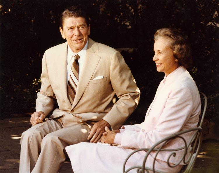 A man in a beige suit and a woman in a light pink suit are seated outdoors, smiling. The setting is a shaded garden or patio with greenery in the background. The man appears to be holding papers in his hand.