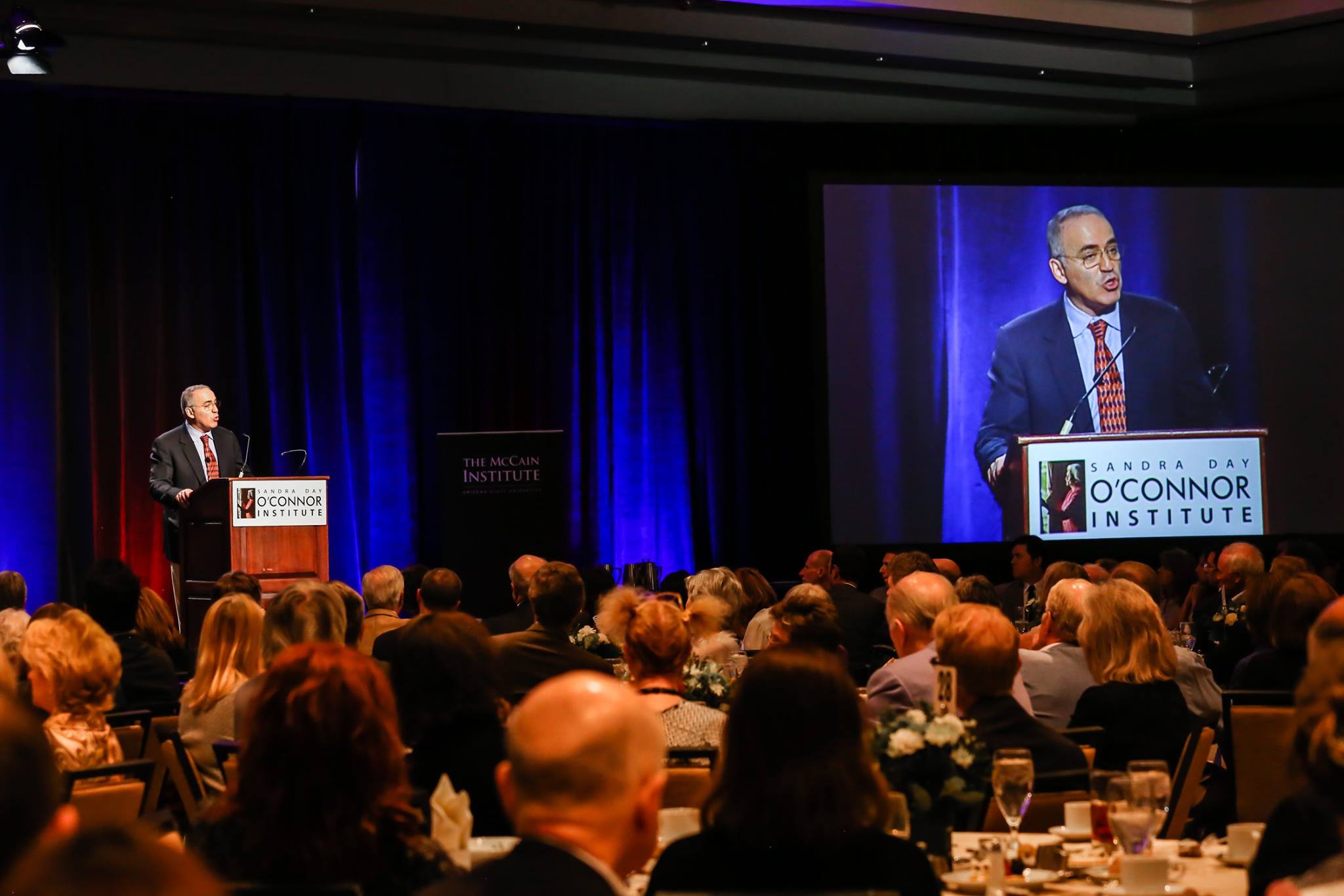 A speaker at a podium addresses a large seated audience in a dimly lit conference room. A projection screen displays the speaker. The event is hosted by the Sandra Day O'Connor Institute and the McCain Institute, as indicated by the signs.