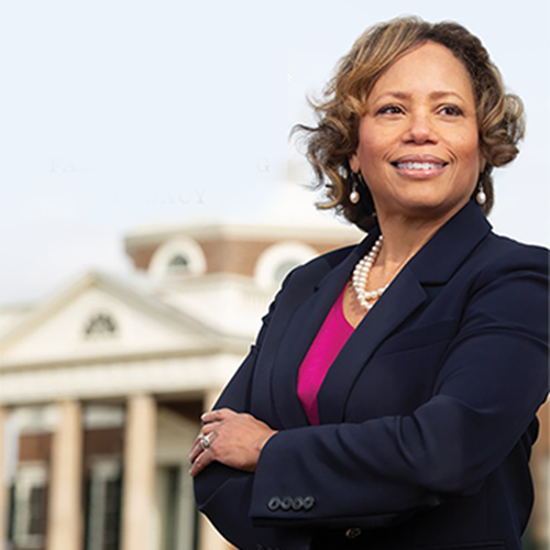 A person in a navy blazer stands confidently outside, with a building with neoclassical architecture and columns in the background. They are smiling and have curly hair.