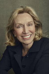 A smiling woman with shoulder-length blonde hair in a black jacket poses against a textured backdrop, reminiscent of a classic dinner scene from history.