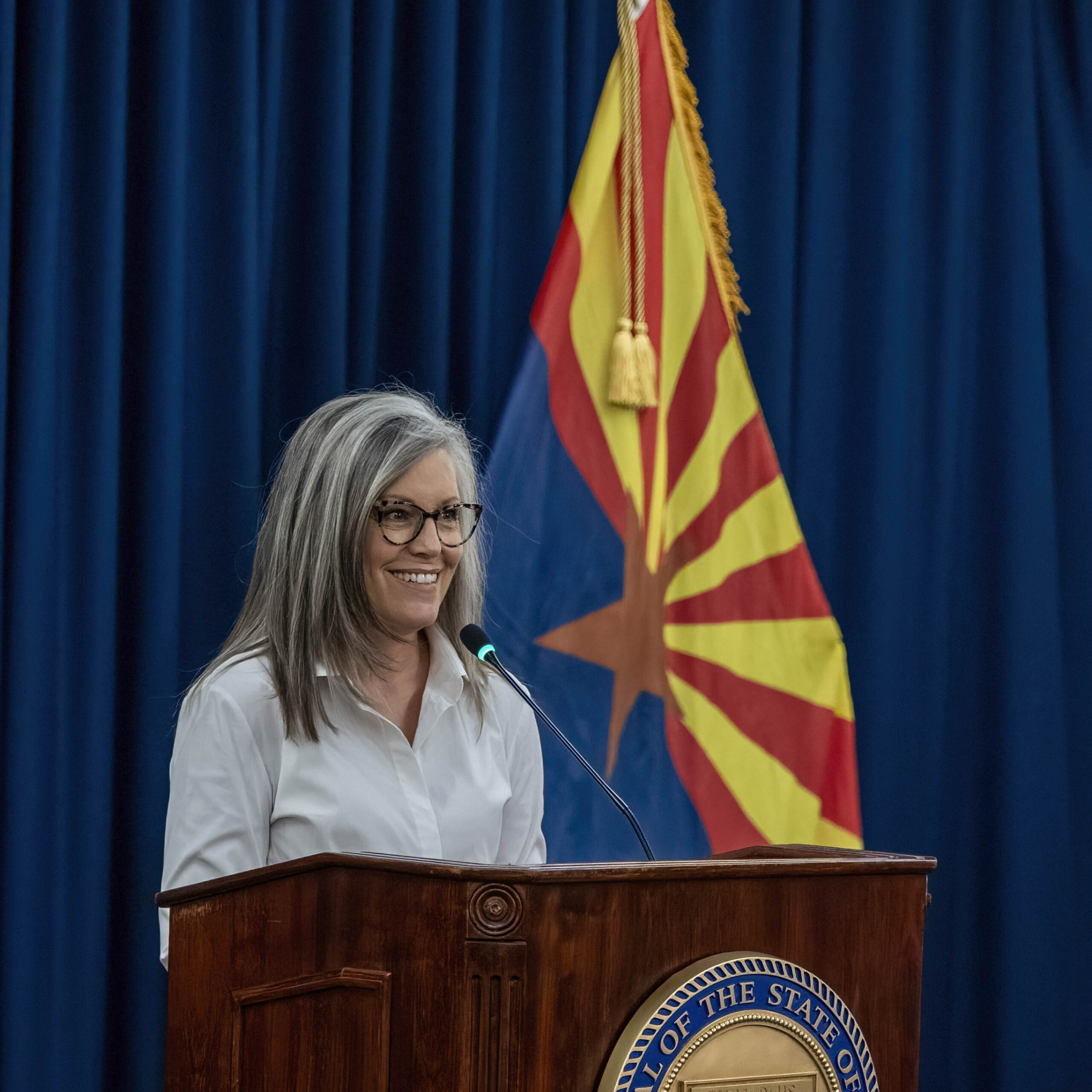 June 10, 2025 Camp SDO A gray-haired woman with glasses stands at a podium, smiling and speaking into a microphone. Behind her is a blue curtain and the Arizona state flag, highlighting her role as an active citizen in the community.