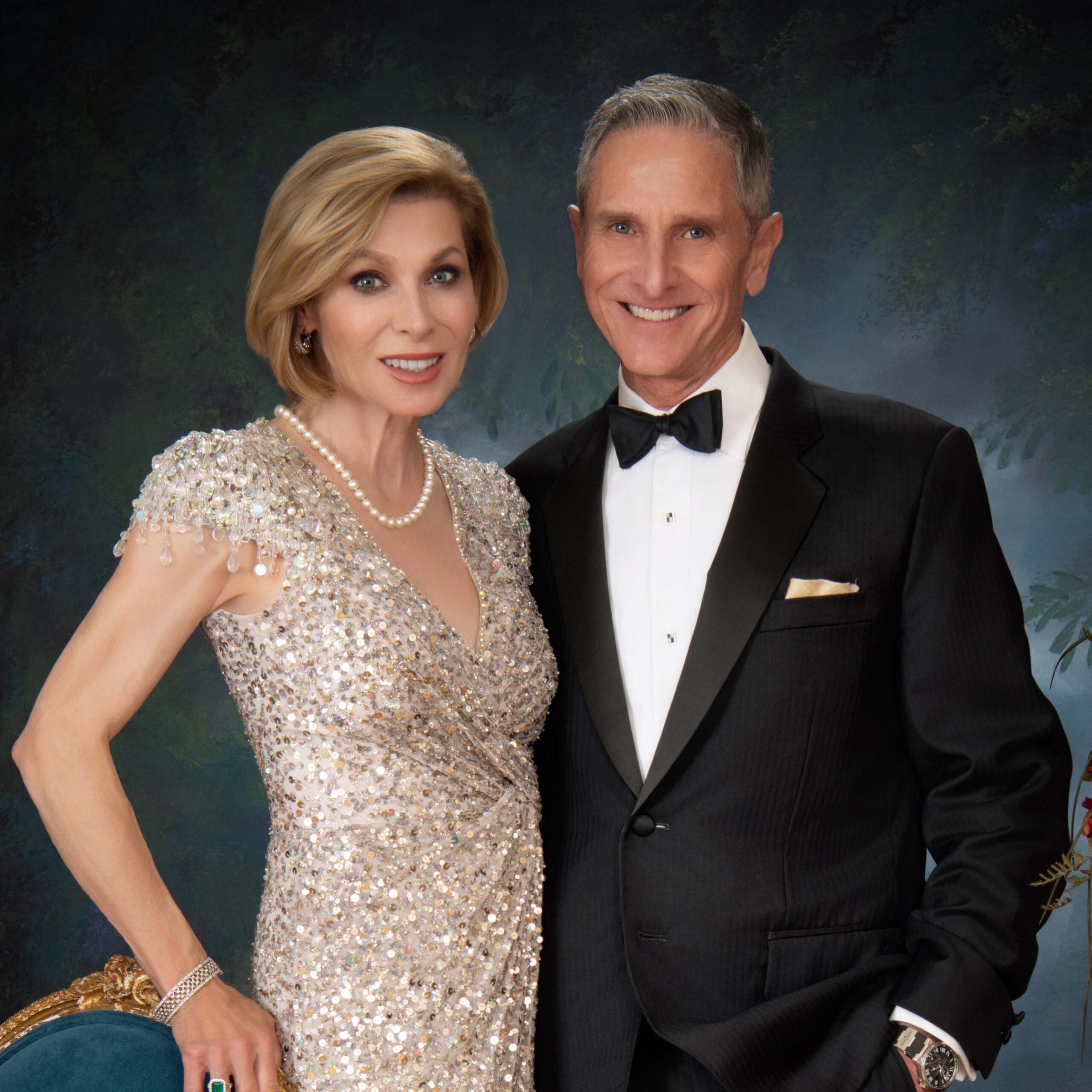 A woman in a sparkling, silver evening gown stands beside a man in a black tuxedo and bow tie. Both are smiling, posing at what appears to be a history dinner, with an elegant painted backdrop adding formal flair.