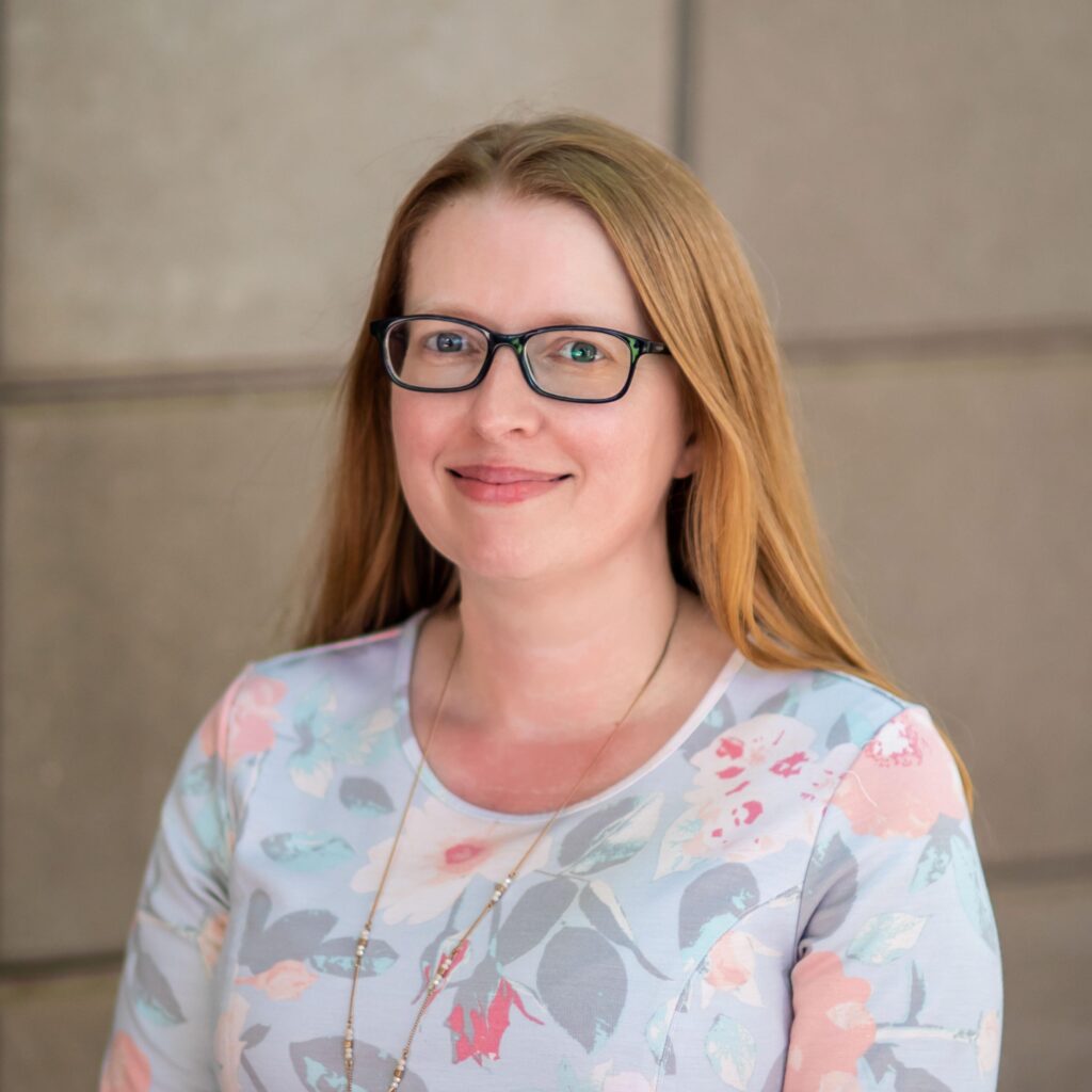 A woman with long blonde hair and glasses smiles at the camera. She is wearing a floral-patterned top and standing against a light-colored, tiled wall.