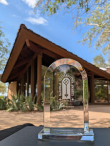 A glass National Preservation Award trophy sits on a table outdoors, with a wooden building and trees in the background under a clear blue sky.