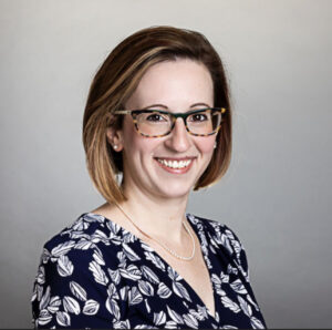 A smiling woman with shoulder-length brown hair, wearing tortoiseshell glasses, a patterned blouse, and a pearl necklace. She poses against a neutral gray background.