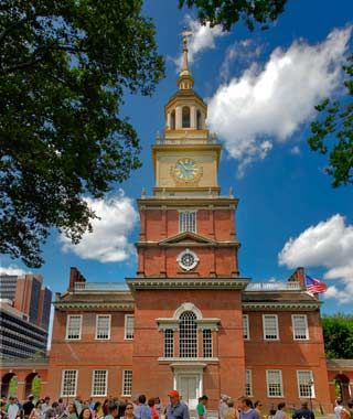 A crowd gathers in front of Independence Hall, where the Constitution was signed, a historic red-brick building with a tall clock tower, under a blue sky with scattered clouds and nearby trees.