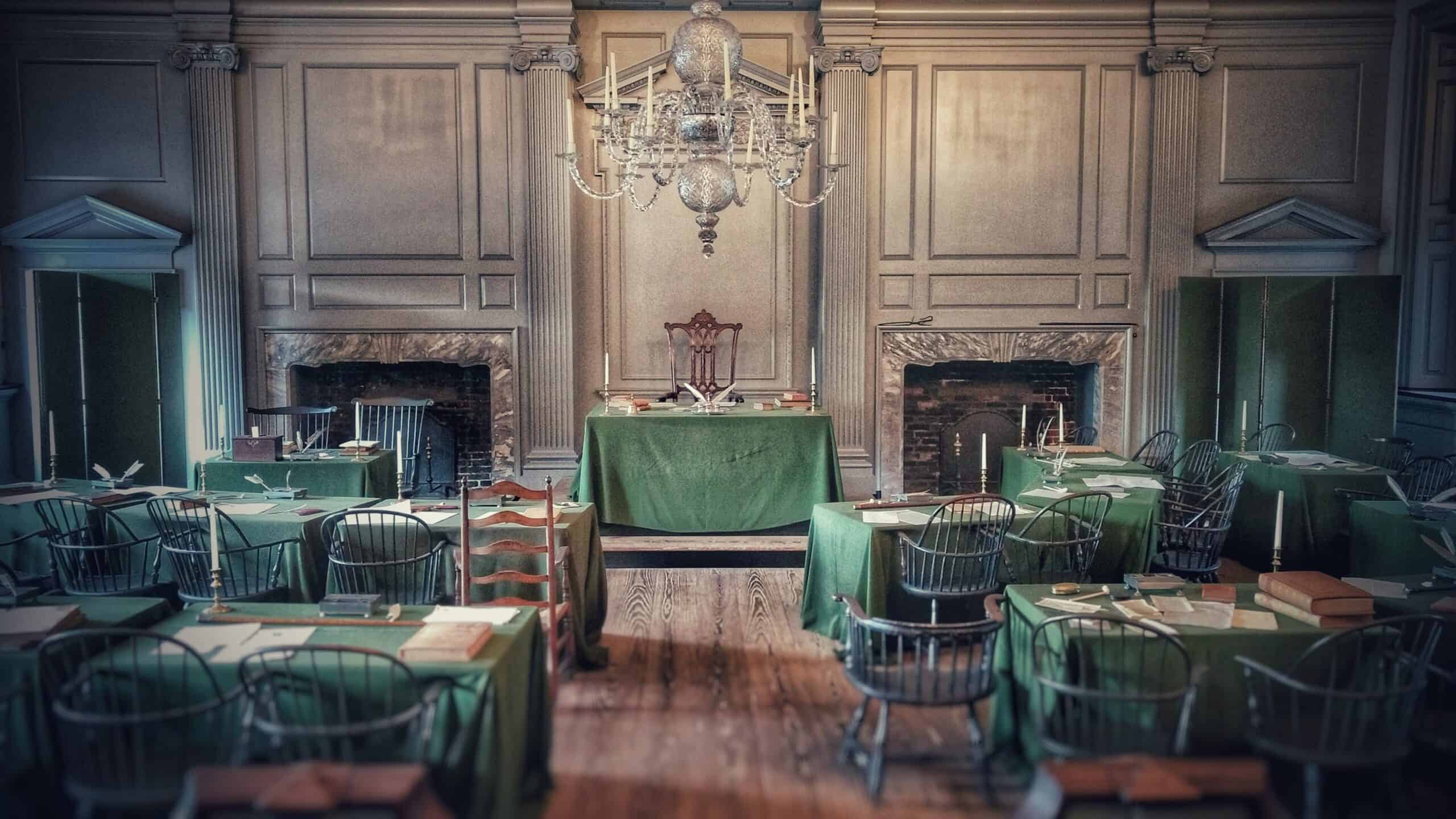 A historic meeting room with wooden floors, green-covered tables, and a grand chandelier—perfect for a Summer of Civics program. Papers and inkwells adorn the colonial-era space, flanked by two fireplaces at the front for a formal atmosphere.