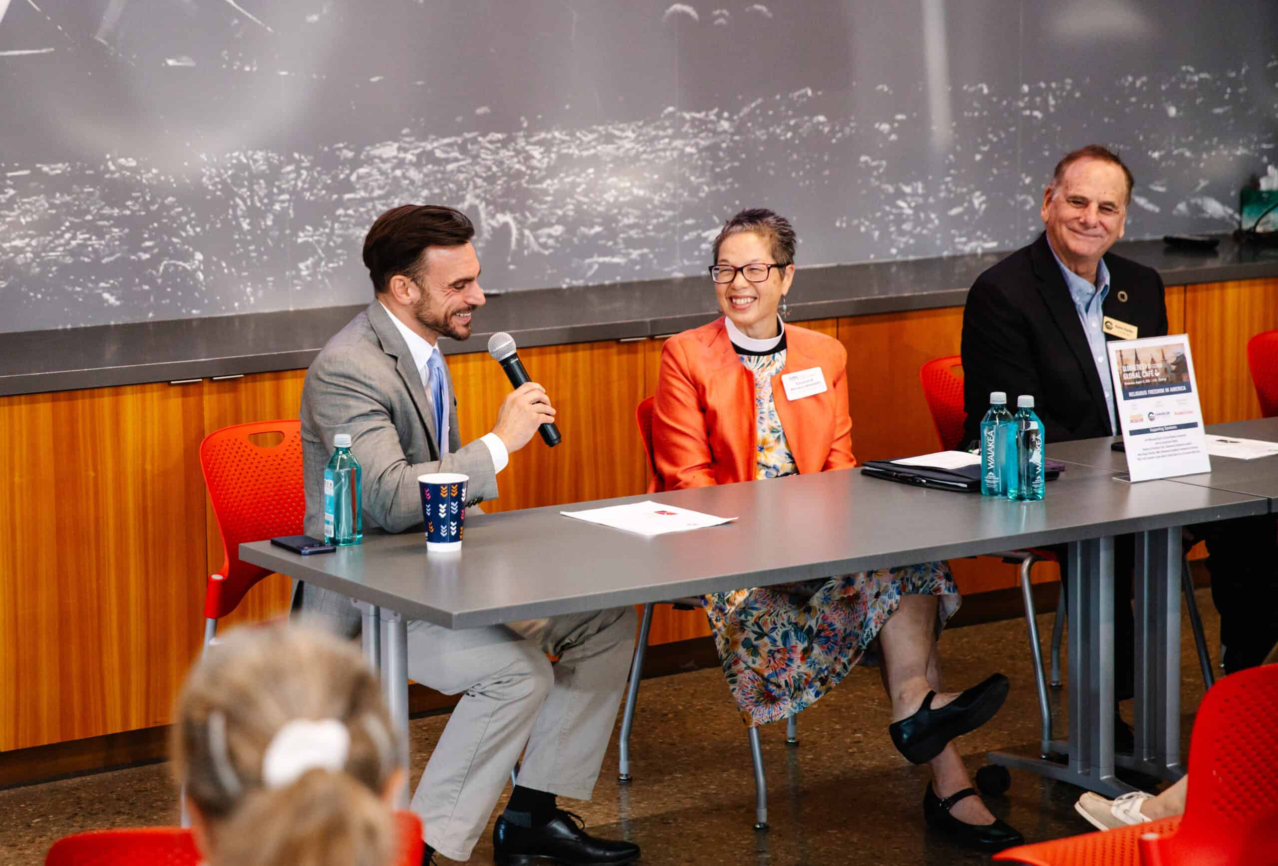 Three people sit at a table on a panel. The man on the left speaks into a microphone, while the woman and man next to him smile. Bottles of water and papers are scattered across the table, including a folder labeled “The Citizen.” Audience members are visible in the foreground.