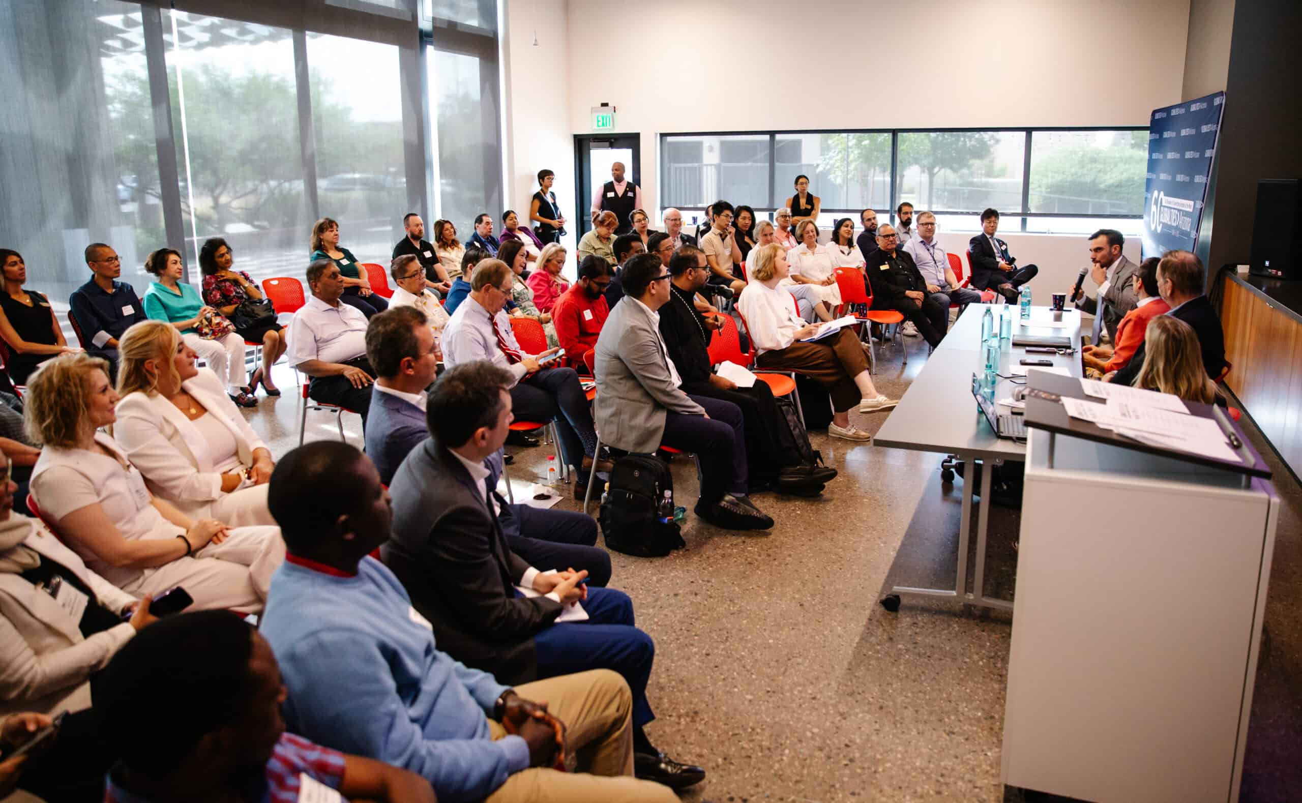 A diverse group of people, including members of The Citizen, sit in rows facing a panel of speakers in a modern, bright conference room with large windows and red chairs.