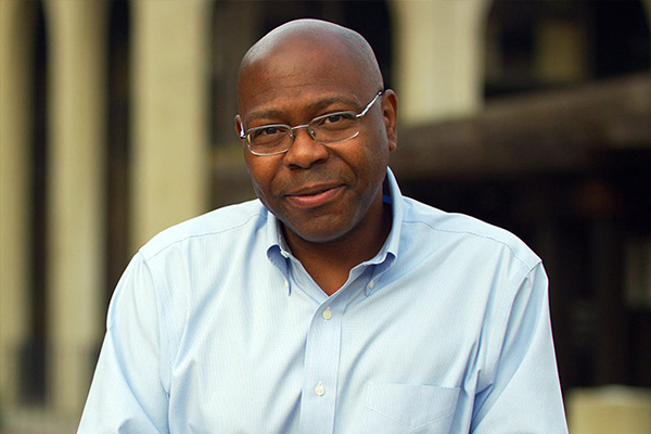 A man in glasses and a light blue shirt is standing outdoors, looking at the camera with a slight smile. There is an architectural structure blurred in the background.