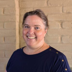 A woman with light skin and brown hair pulled back smiles at the camera. She is wearing a navy blue top with gold buttons and stands in front of a beige brick wall.