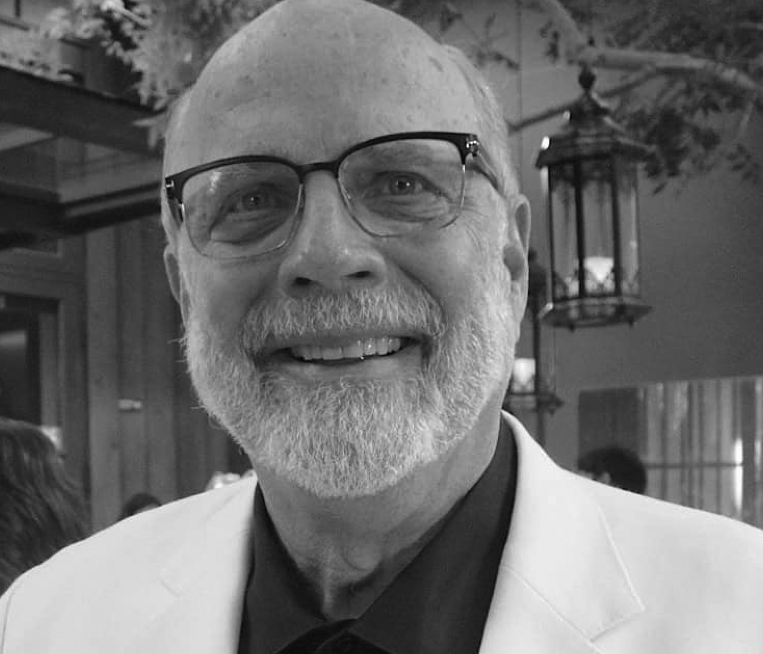 Older man with glasses, a white beard, and a friendly smile, wearing a light-colored blazer and dark shirt. The background includes hanging lanterns and blurred figures. The photo is in black and white.