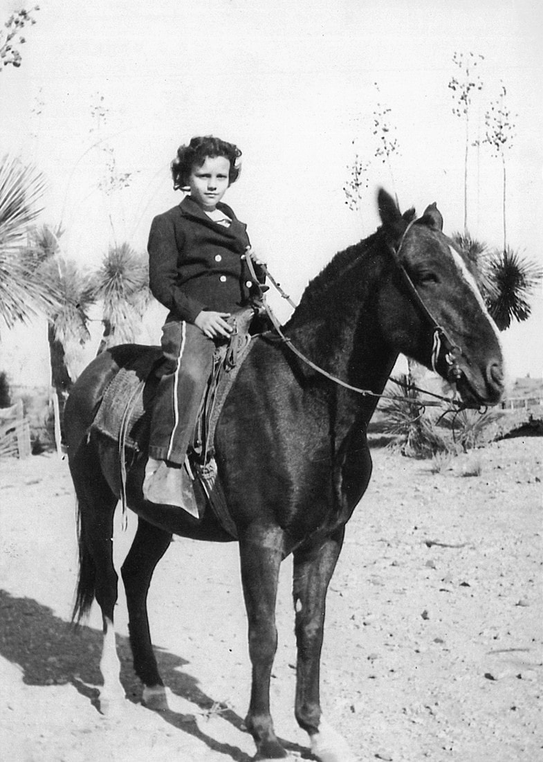 A child wearing a suit and boots sits on a dark horse in a sandy outdoor area with desert plants and trees in the background. The old black-and-white photo evokes a timeless sense of justice and quiet determination.