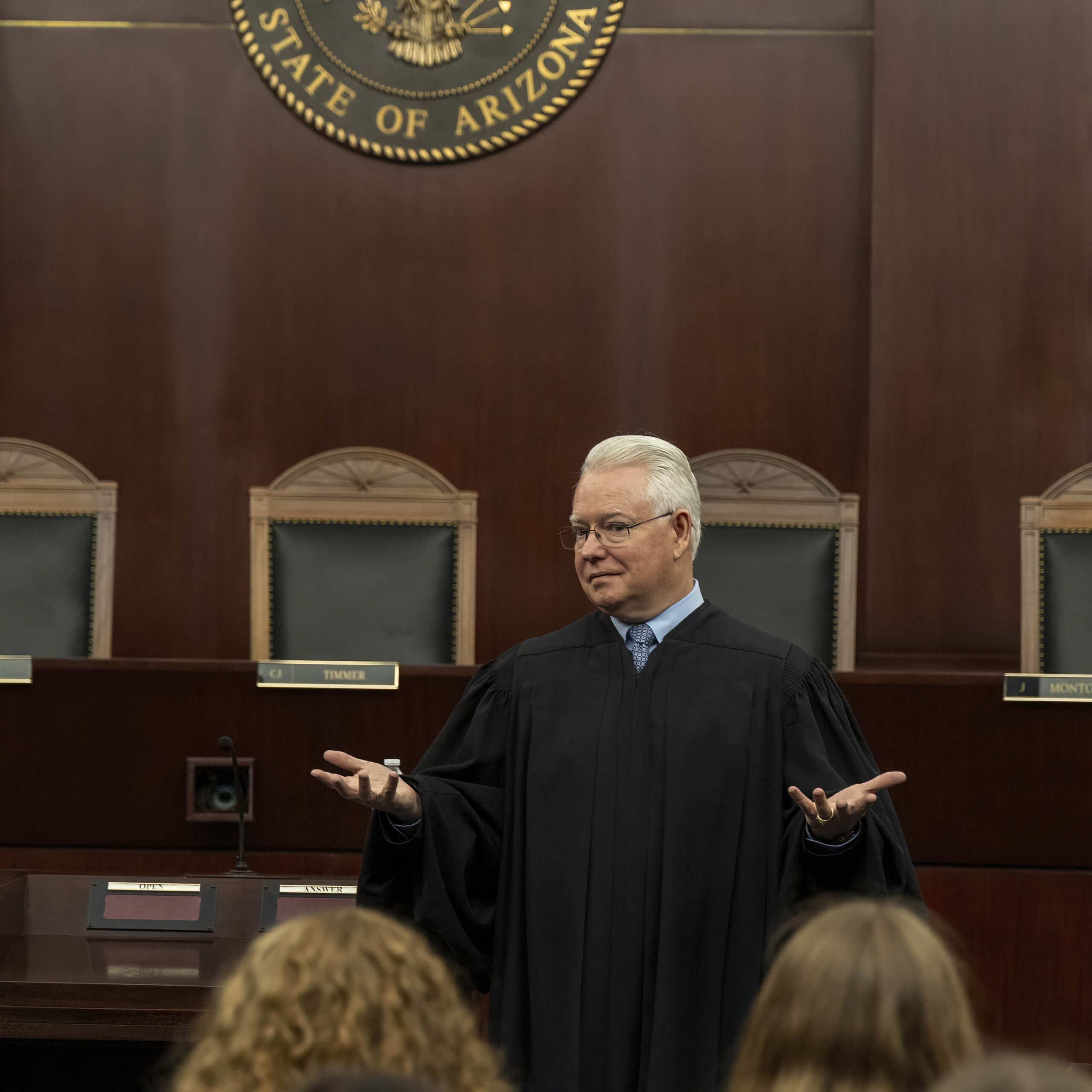 June 9, 2025 Camp SDO A judge in a black robe stands before empty chairs and a wooden bench in a courtroom, gesturing with open hands while addressing citizens seated in the foreground. The Arizona state seal is visible behind him.