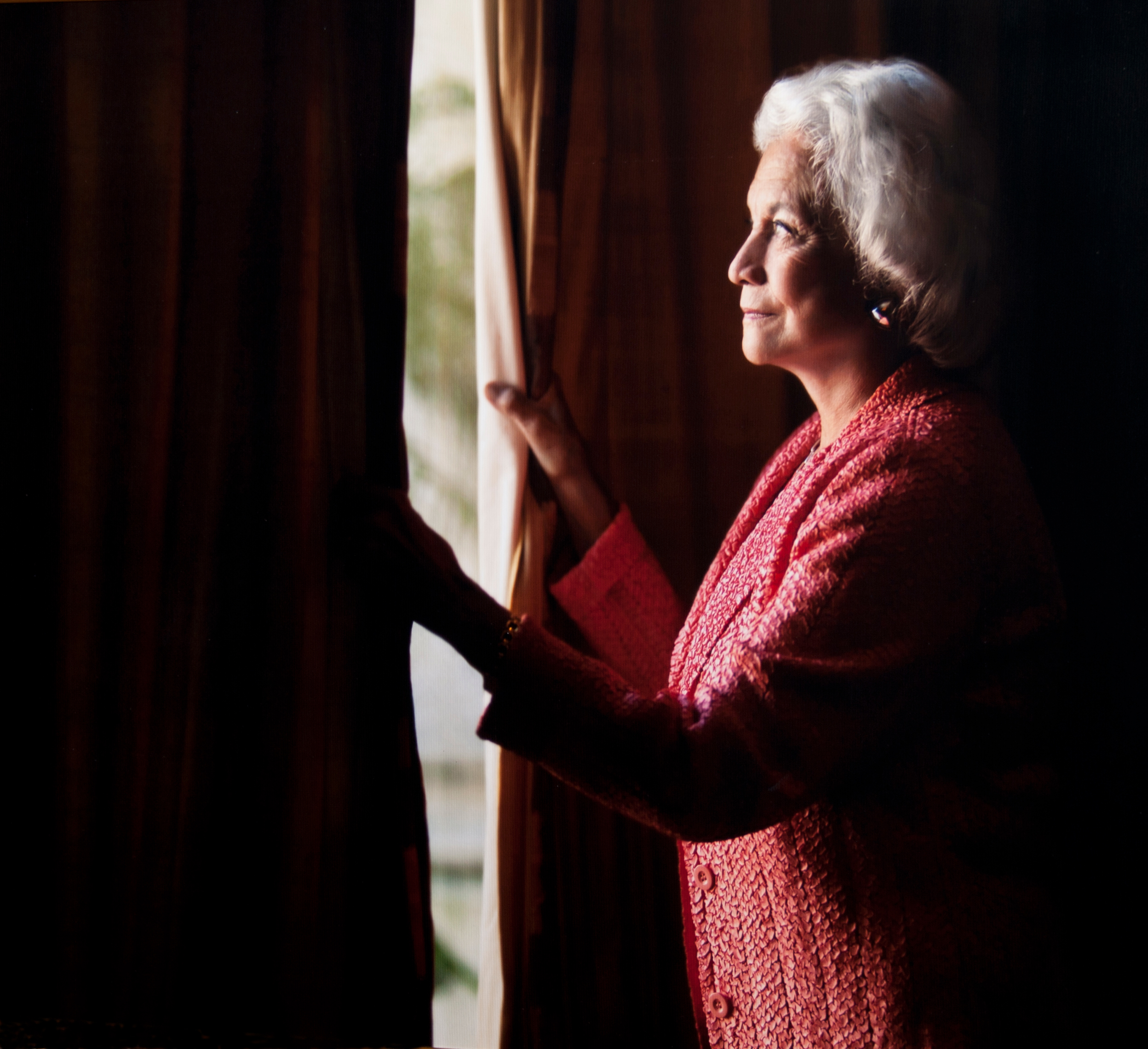 An older woman with white hair, wearing a textured pink jacket, stands by a window. She gently holds the curtains open, gazing outside thoughtfully. The background is softly blurred, emphasizing her profile.