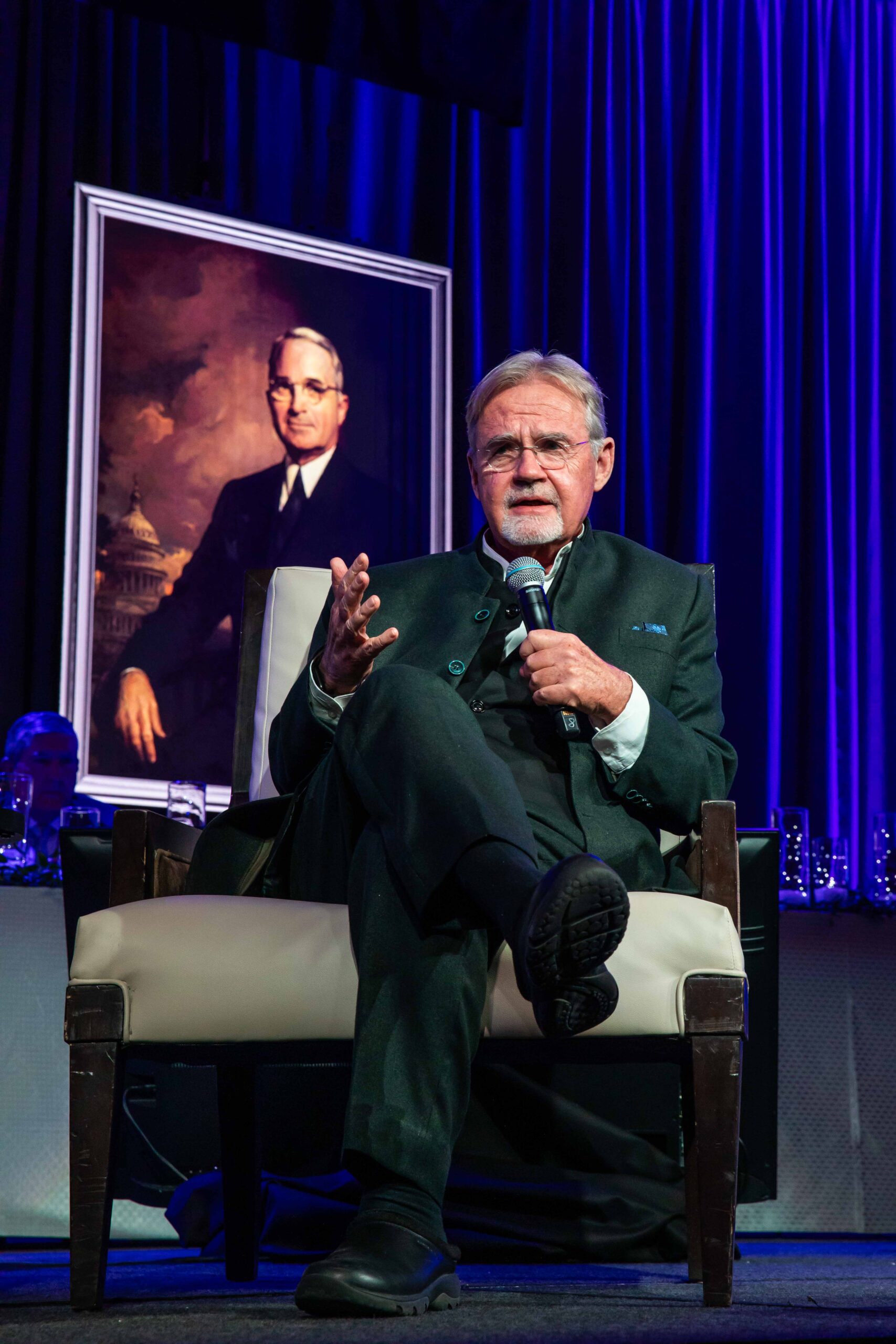 An older man with a beard sits in a chair, speaking into a microphone. He wears a dark suit. Behind him is a large portrait of another man in a suit. The background is a deep blue curtain.