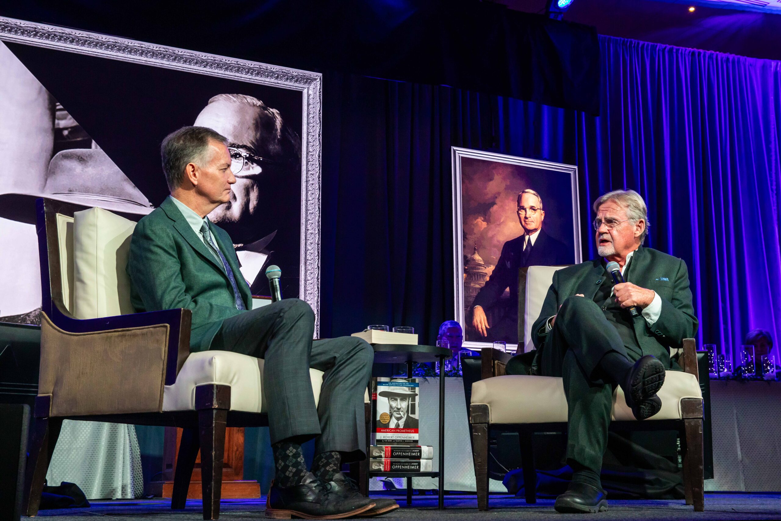 Two men in suits sit on a stage with microphones, engaged in conversation. Behind them are two large framed portraits of a distinguished older man. The setting suggests a formal event or discussion panel.