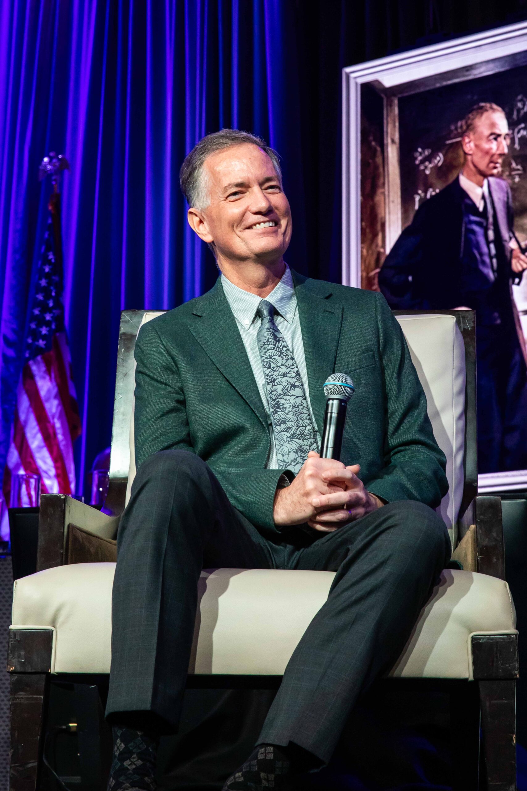 A person in a green suit sits on a chair, holding a microphone, smiling. In the background, there is a portrait and an American flag. Blue curtains and stage lighting create a formal setting.