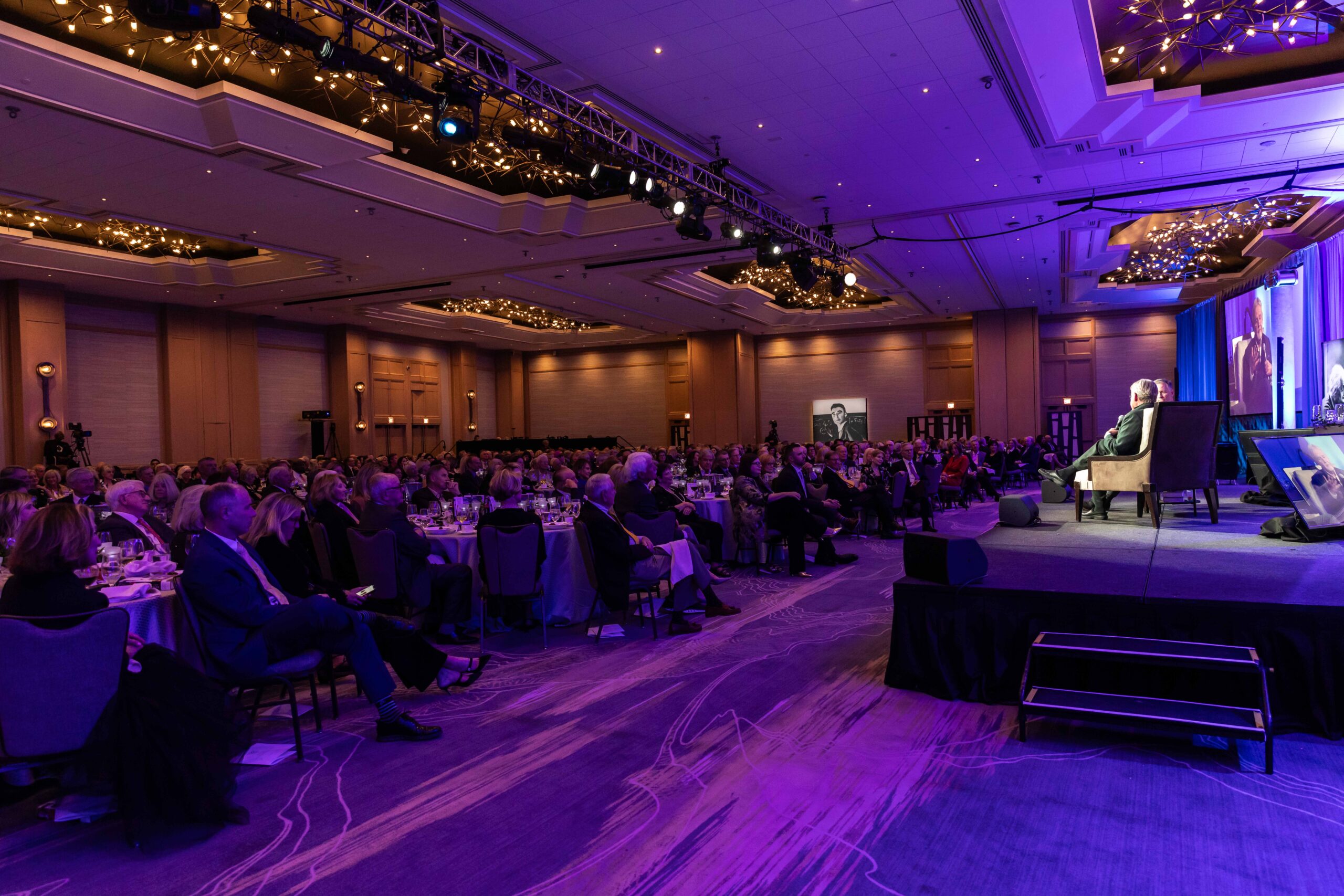 Oppenheimer Truman Crowd A speaker stands on stage in a large, elegantly lit auditorium, urging attendees to donate generously. The room is illuminated with purple lighting, creating an atmosphere of hope as the audience in formal attire listens intently.