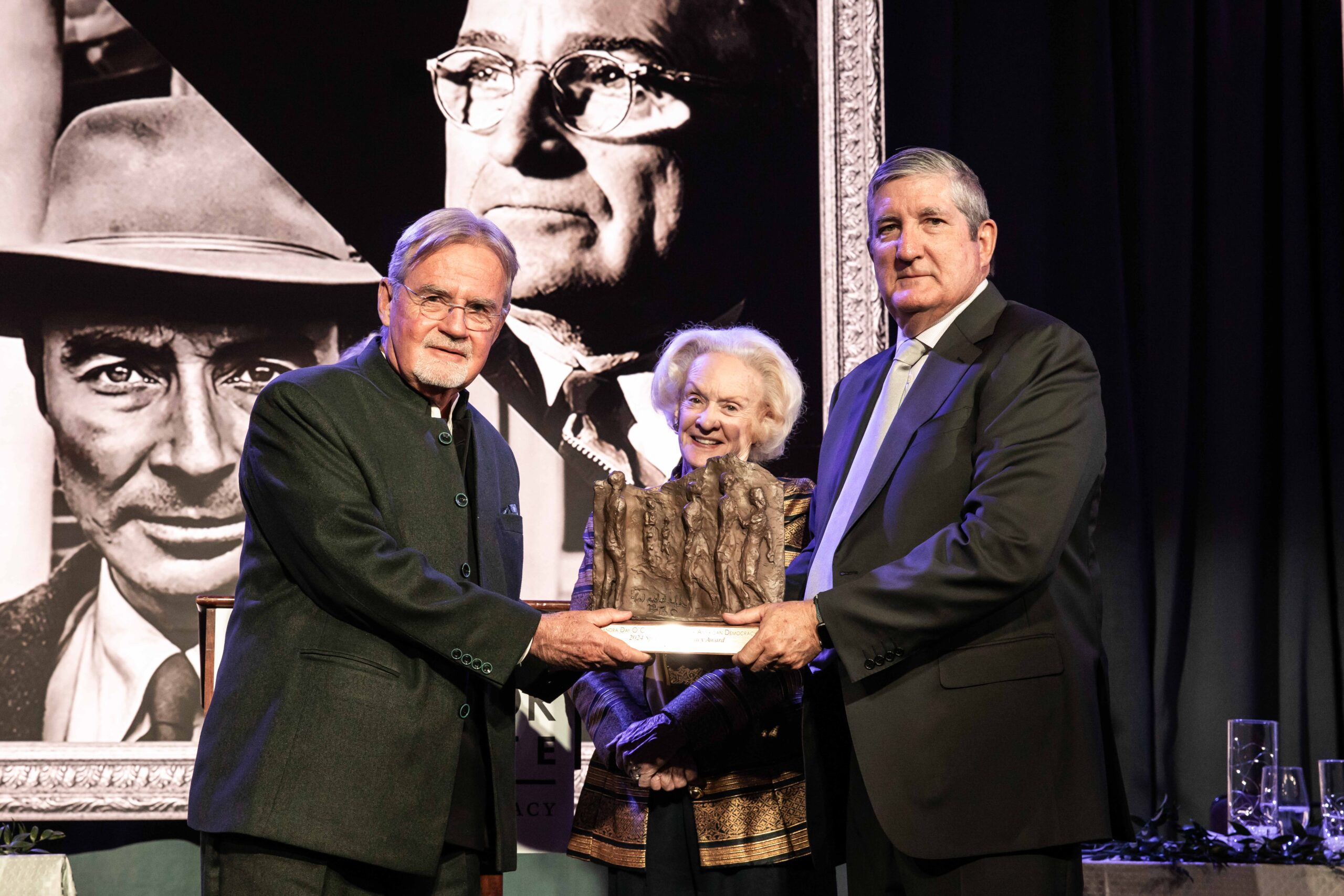 Three older adults stand on a stage, holding a sculpture. Two men and a woman, all in formal attire. Behind them is a large black-and-white photograph of an older man. The atmosphere suggests a formal award ceremony.