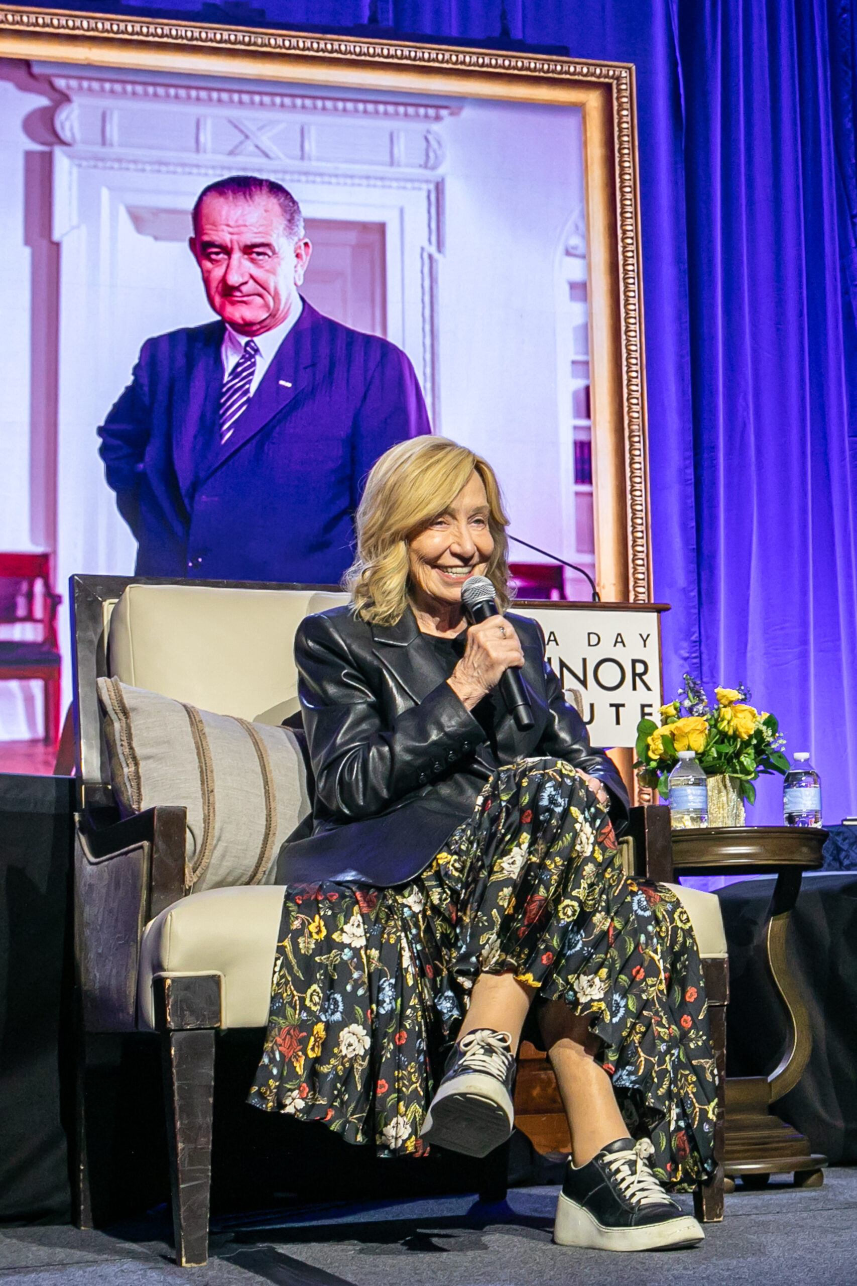 A woman sits in an armchair on stage, smiling and holding a microphone. She wears a floral skirt, leather jacket, and sneakers. Behind her is a large framed portrait of a man in a suit—an atmosphere fit for Doris Kearns Goodwin.