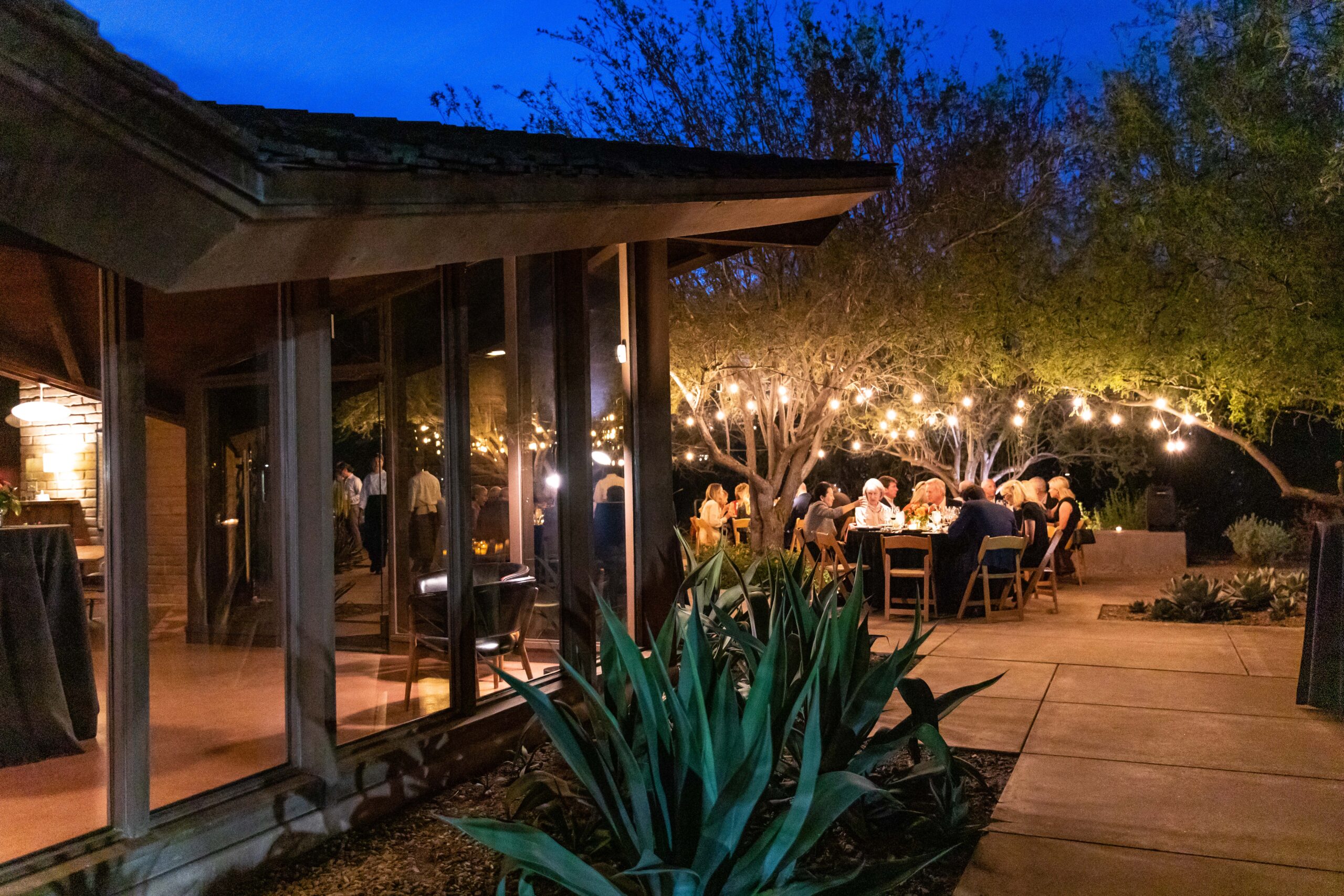 Outdoor evening gathering with people seated at tables under string lights, surrounded by trees and greenery. A modern building with large windows is on the left. The scene conveys a warm and festive atmosphere.