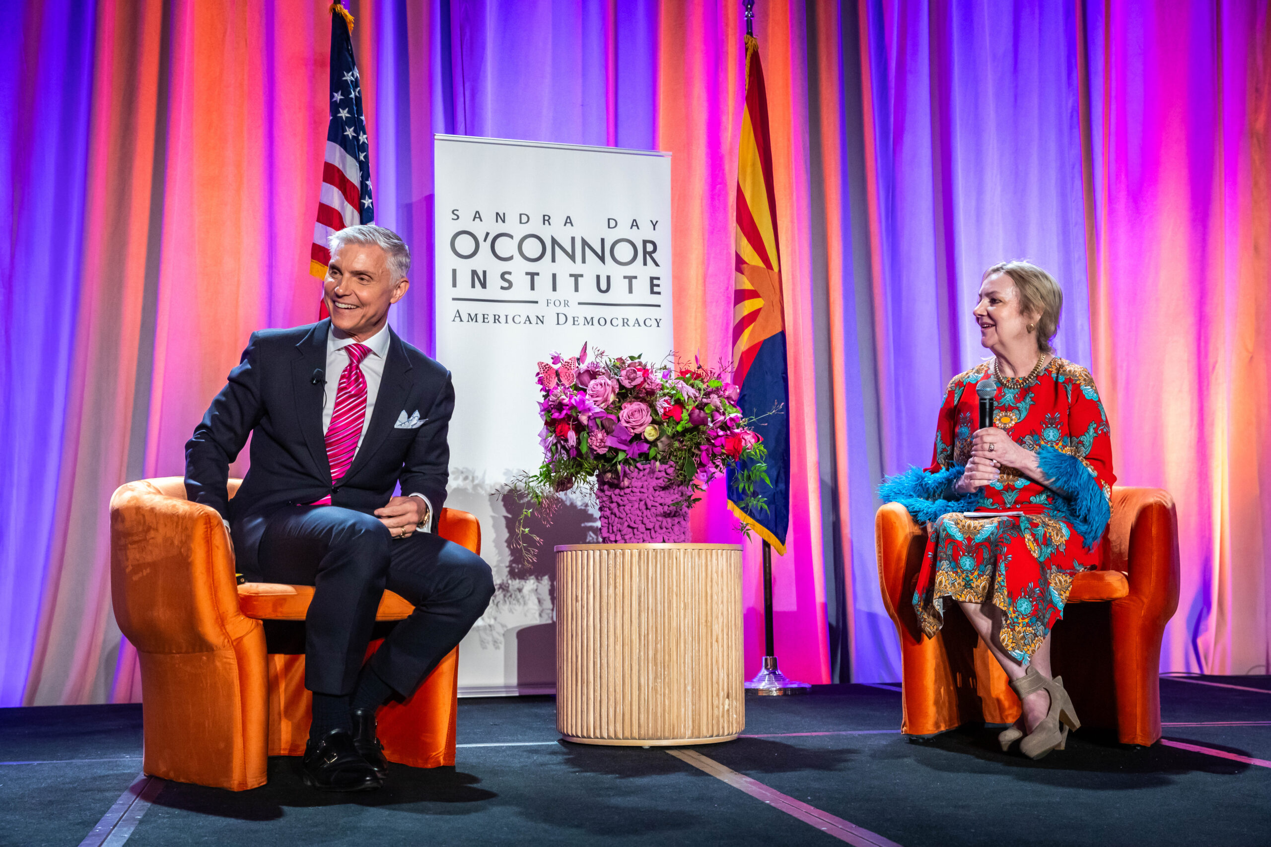 A man in a suit and a woman in a colorful dress sit in orange chairs on a stage. Behind them are banners, an American flag, and floral arrangements. The backdrop features purple and orange lighting. They are engaged in conversation, smiling.